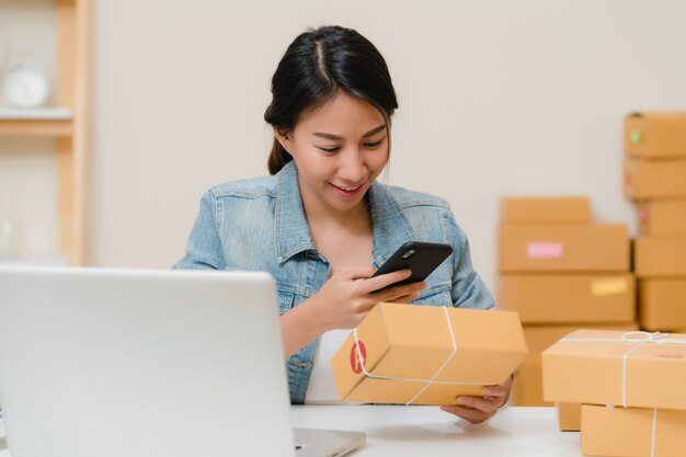 Dueño joven asiático elegante hermoso de la mujer de negocios del empresario de la PYME que comprueba el producto en el código común del qr de la exploración que trabaja en casa.