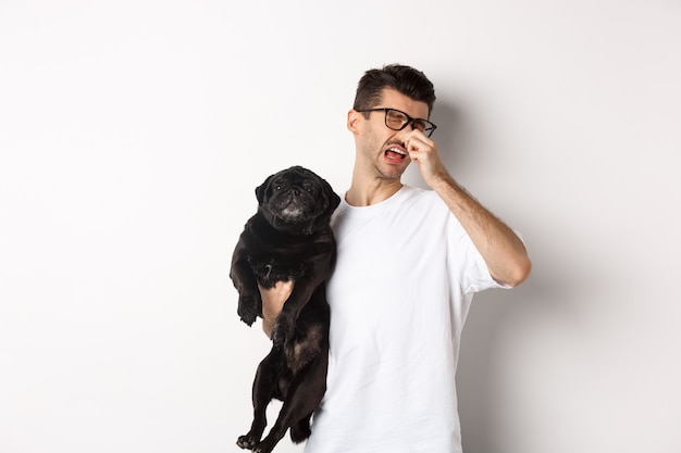 Dueño del perro entrecerrar los ojos y cerrar la nariz por un hedor repugnante, sosteniendo pug con olor a cama, de pie sobre fondo blanco.