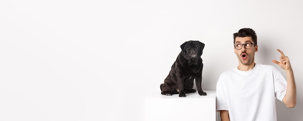 Dueño de un perro macho feliz sentado cerca de un lindo pug negro mirando a la mascota con la cara asombrada y sorprendida blanca