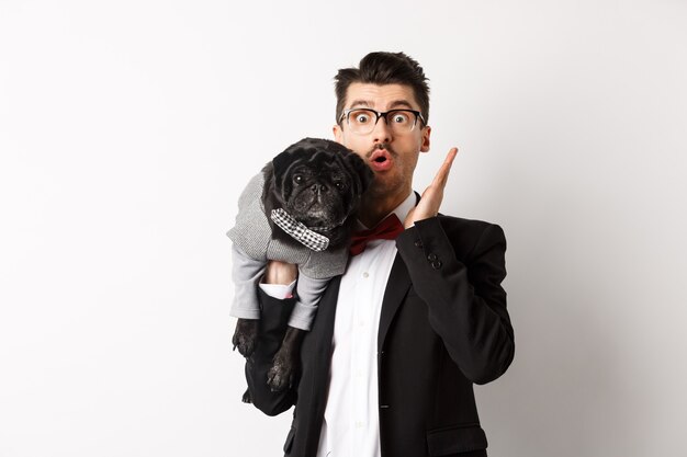 Dueño de perro sorprendido y asombrado en traje mirando a la cámara, sosteniendo lindo pug negro en el hombro, traje de mascota, posando sobre fondo blanco