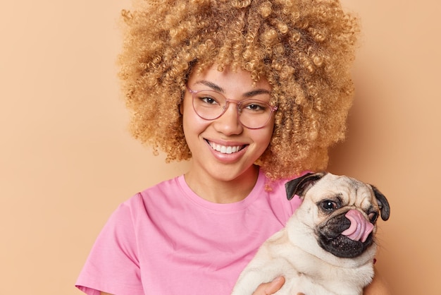 El dueño positivo de una mascota sonríe alegremente vestido con una camiseta rosa informal posa con un perro pug que va a caminar y se siente contento aislado sobre un fondo marrón A la modelo femenina de pelo rizado le gustan los animales domésticos
