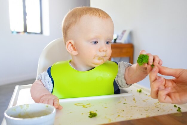 Dulce bebé en babero tomando una pieza de brócoli de mamá