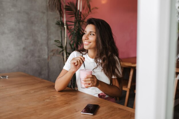 Dulce chica bronceada está revolviendo su batido de leche Mujer sentada en el café en la mesa de madera