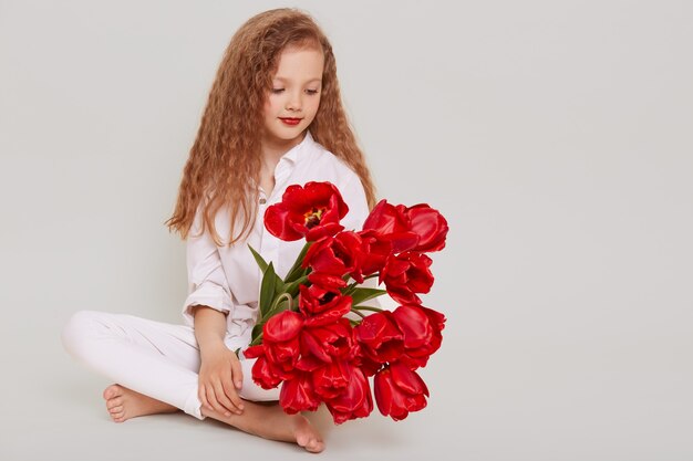 Dulce chica rubia con cabello ondulado sentada en el piso y mirando tulipanes rojos en sus manos, con expresión soñadora
