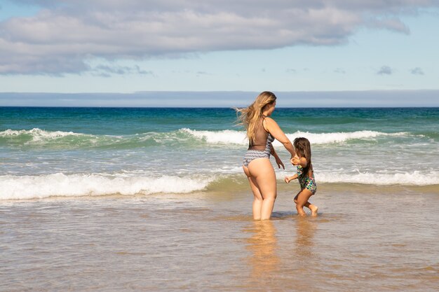 Dulce joven madre e hija de pie hasta los tobillos en el agua de mar, pasando el tiempo libre en la playa en el océano