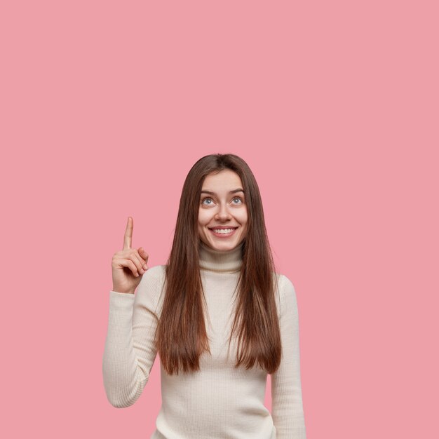 Dulce linda mujer joven con puntos de pelo largo y oscuro con los dedos índices hacia arriba, mira al techo, vestida con ropa casual