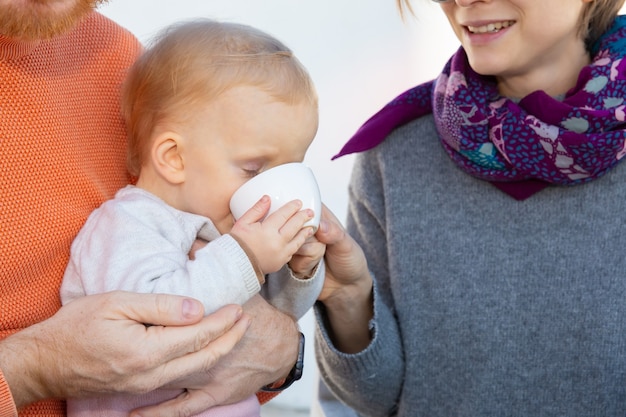 Dulce niña bebiendo de la taza blanca mientras está sentado en el regazo de los papás