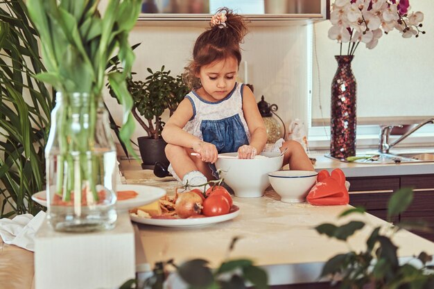 Dulce niña linda aprende a cocinar una comida en la cocina mientras se sienta en una encimera.