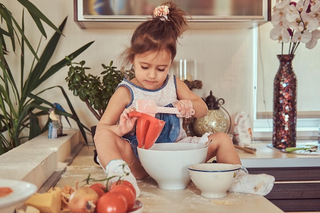 Dulce niña linda aprende a cocinar una comida en la cocina mientras se sienta en una encimera.