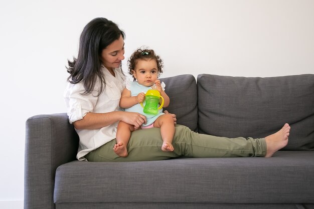 Dulce niña sentada en el regazo de las mamás y bebiendo agua de la botella. Copie el espacio. Concepto de paternidad e infancia