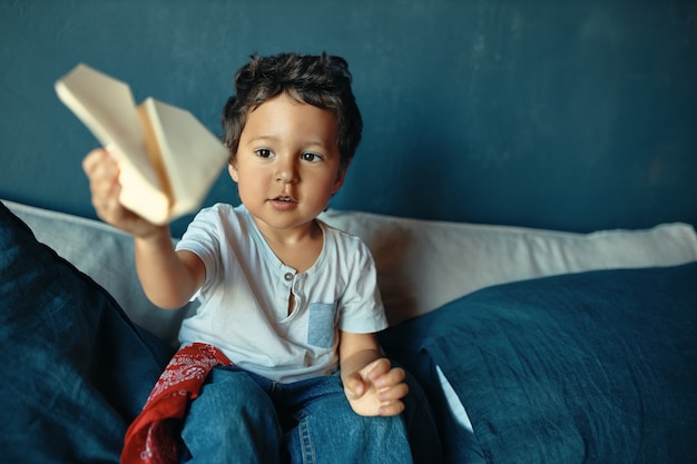 Dulce niño gordito de piel oscura sentado en la cama, jugando juegos activos, lanzando un avión de papel, con una expresión facial emocionada.