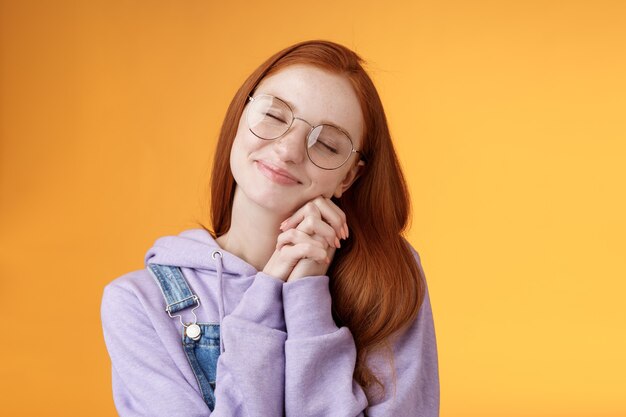 Dulce tierna feliz romántica pelirroja novia ojos cerrados sonriendo imágenes de ensueño escena encantadora soñar despierto prensa palmas mejillas tonto recordando bonitos recuerdos, de pie fondo naranja relajado