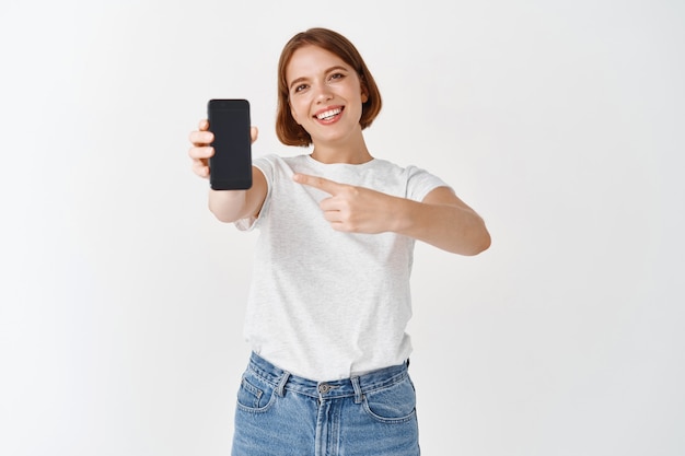 Echale un vistazo. Sonriente mujer hermosa en ropa casual, apuntando a la pantalla vacía del teléfono inteligente, mostrando tienda o aplicación en línea, pared blanca