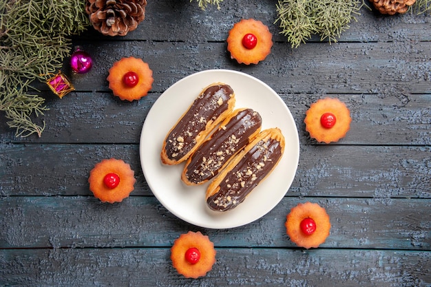Foto gratuita eclairs de chocolate de vista superior en plato ovalado blanco rodeado de cupcakes ramas de abeto juguetes de navidad en suelo de madera oscura con espacio libre