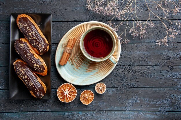 Eclairs de chocolate de vista superior en un plato rectangular y una taza de té de limones secos y canela en la mesa de madera oscura con espacio libre