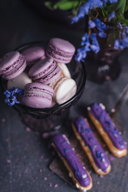 Eclairs morados en espátula con macarrones en un recipiente de vidrio cerca del jarrón