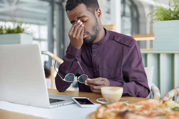 Economista masculino profesional con exceso de trabajo, sostiene gafas, está cansado de trabajar en la computadora portátil muchas horas, tiene dolor de cabeza después de un día de trabajo cansado