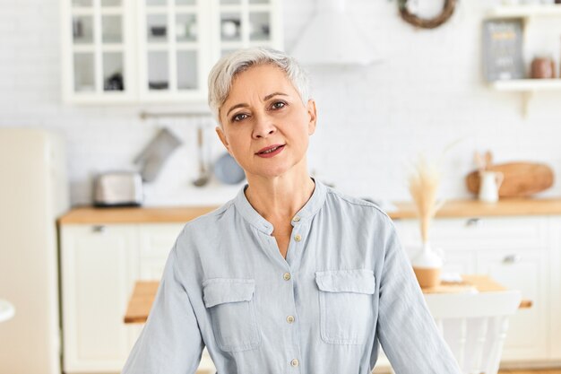 Edad, madurez, domesticidad y estilo de vida. Filmación en interiores de pensionista vestida de manera informal con aspecto cansado, exhalando después de hacer todas las tareas del hogar, posando contra el interior de la cocina borrosa