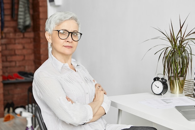 Edad, madurez, profesión y empleo. Filmación en interiores de una mujer independiente de cincuenta años de edad, seria y segura que trabaja en la oficina en casa mientras está de jubilación, usando la computadora portátil, cruzando los brazos sobre el pecho