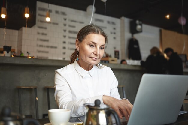 Edad, ocupación, autónomo y concepto de trabajo remoto. Hermosa mujer madura europea de pelo gary freelancer trabajando en un proyecto a distancia, utilizando una conexión a Internet de alta velocidad en un portátil en la cafetería.