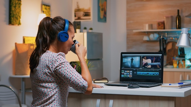 Editor de video de mujer con auriculares trabajando con imágenes y sonido sentado en la cocina de casa. Mujer camarógrafo edición de montaje de película de audio en portátil profesional sentado en el escritorio a medianoche