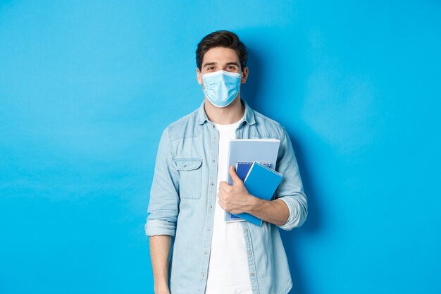 Educación, covid-19 y distanciamiento social. Estudiante de chico en máscara médica mirando feliz, sosteniendo cuadernos, de pie sobre fondo azul.