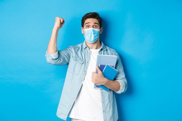 Educación, covid-19 y distanciamiento social. Estudiante masculino emocionado en máscara médica triunfando, levantando el puño y sosteniendo cuadernos, de pie sobre fondo azul.