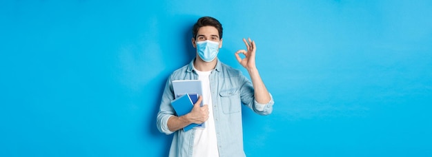 Foto gratuita educación covid y estudiante de distanciamiento social con máscara médica que parece feliz sosteniendo cuadernos sh