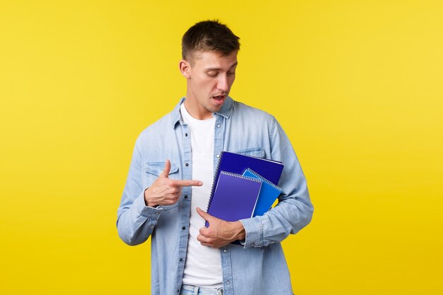 Educación, cursos y concepto universitario. Apuesto joven estudiante varón apuntando con el dedo a cuadernos y material de estudio, preparar la tarea para la asignación universitaria, fondo amarillo.