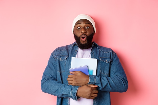Educación. Estudiante adulto afroamericano emocionado lleva cuadernos, mirando a la cámara asombrado, de pie sobre fondo rosa.