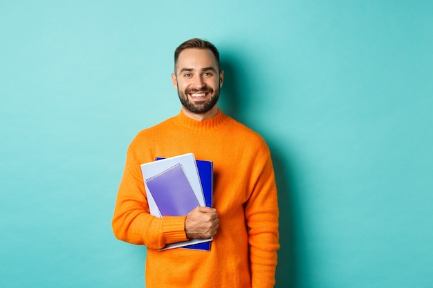 Educación. Hombre barbudo sonriente sosteniendo cuadernos y sonriendo, yendo a cursos, de pie sobre una pared turquesa clara
