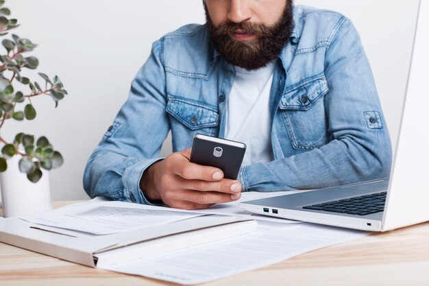 Un efecto cinematográfico. Retrato de hombre barbudo en jean camisa hoding smartphone en su mano mientras trabajaba con documentos en la oficina con acogedor interior. Retrato recortado del exitoso hombre de negocios mediante dispositivo móvil.