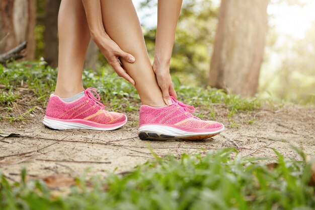 Ejecución de lesiones deportivas. Corredor de la atleta femenina con zapatillas rosas tocando su tobillo torcido o torcido mientras trota o corre al aire libre.