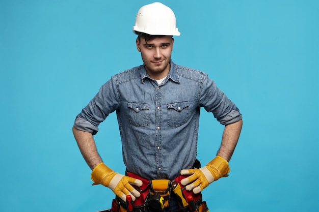 Electricista masculino joven experto profesional en casco protector y guantes tomados de la mano en la cintura