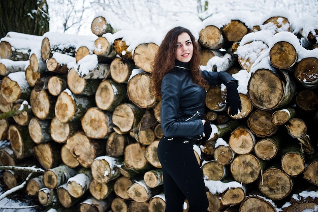Elegancia chica rizada con chaqueta de cuero en el parque forestal nevado en invierno contra tocones