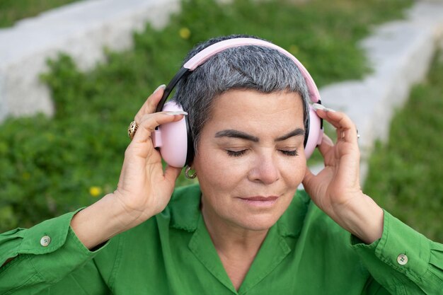 Elegante anciana escuchando música en el parque. Modelo femenino con cabello gris corto en ropa brillante y grandes auriculares. Ocio, actividad, concepto de tecnología.