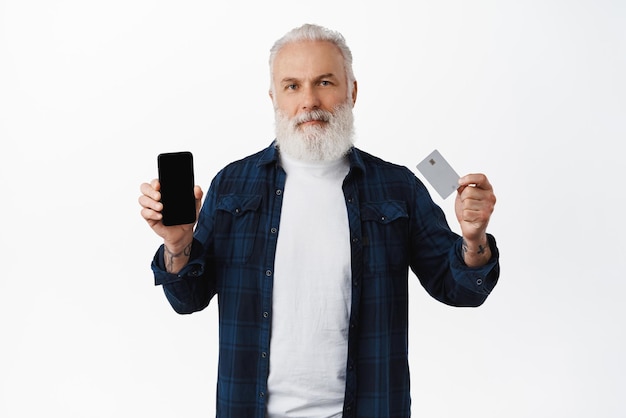 Elegante anciano con barba que muestra la pantalla del teléfono inteligente y la tarjeta de crédito que recomienda la aplicación de compras o la función bancaria sobre fondo blanco