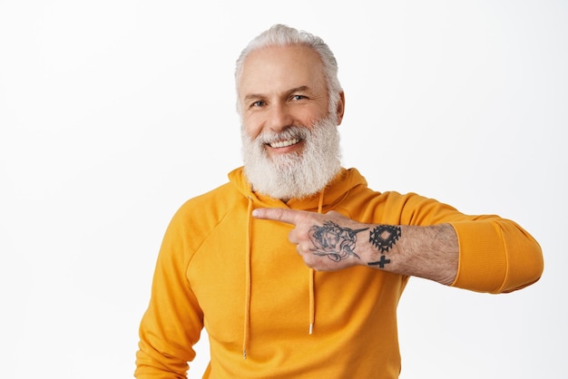 Elegante anciano con tatuajes apuntando con el dedo a la izquierda riendo y sonriendo feliz mostrando el logotipo del anuncio a un lado en el espacio de la copia de pie en una sudadera con capucha naranja sobre fondo blanco