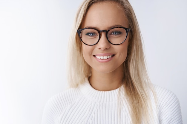 Elegante y atractiva empresaria joven en gafas sonriendo encantada, satisfecha con el nuevo marco de gafas mirando amigable y feliz a la cámara alcanzando el éxito listo para compartir consejos sobre pared blanca.