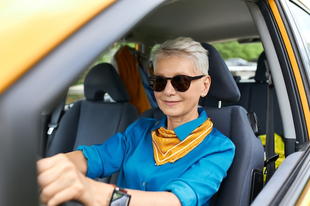 Elegante y atractiva mujer ocupada de mediana edad con gafas de sol y reloj de pulsera yendo de compras, conduciendo su coche nuevo, con mirada de confianza