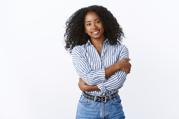 Foto gratuita elegante y atractiva tierna mujer joven afroamericana de pelo rizado abrazando a sí misma mostrando una mujer fuerte y suave al mismo tiempo, sonriendo abrazos de ensueño, con camisa de cuello, jeans, pared blanca