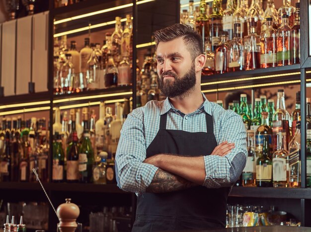 Elegante barman brutal con una camisa y un delantal de pie con los brazos cruzados en el fondo del mostrador del bar.