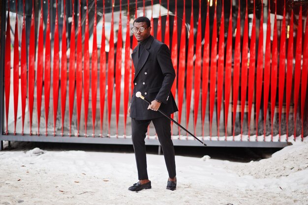 Elegante caballero afroamericano en elegante chaqueta negra con bastón retro como matraz de caña o bastón bastón con mango de bola de diamante dorado Hombre afro rico de moda contra la pared roja