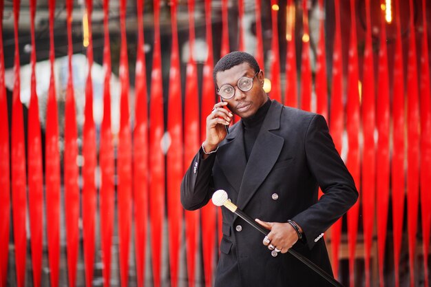Elegante caballero afroamericano en elegante chaqueta negra con bastón retro como matraz de caña o bastón para beber Hombre afro rico de moda contra la pared roja hablando por teléfono móvil