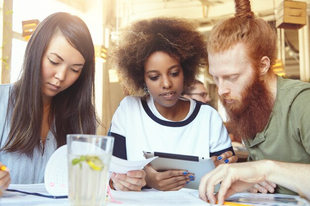 Elegante chica africana con anillo en la nariz usando tableta digital junto con su colega hipster con barba gruesa mientras una mujer asiática seria haciendo papeleo durante la sesión de lluvia de ideas en el espacio de coworking