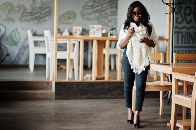 Elegante chica afroamericana con gafas de sol posada en un café moderno y comiendo helado