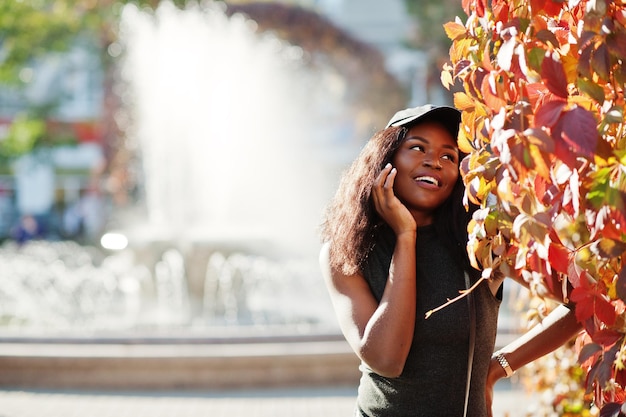 Elegante chica afroamericana con gorra posada en el soleado día de otoño contra hojas rojas Mujer modelo de África