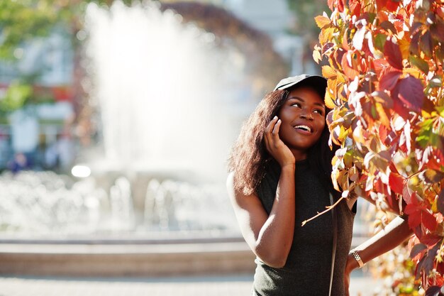 Elegante chica afroamericana con gorra posada en el soleado día de otoño contra hojas rojas Mujer modelo de África