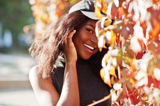 Elegante chica afroamericana con gorra posada en el soleado día de otoño contra hojas rojas Mujer modelo de África