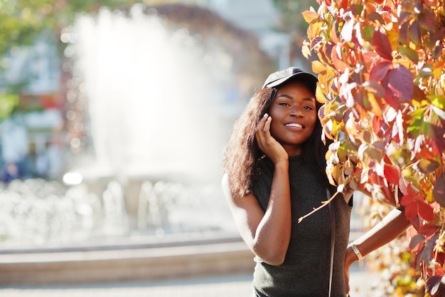 Elegante chica afroamericana con gorra posada en el soleado día de otoño contra hojas rojas Mujer modelo de África